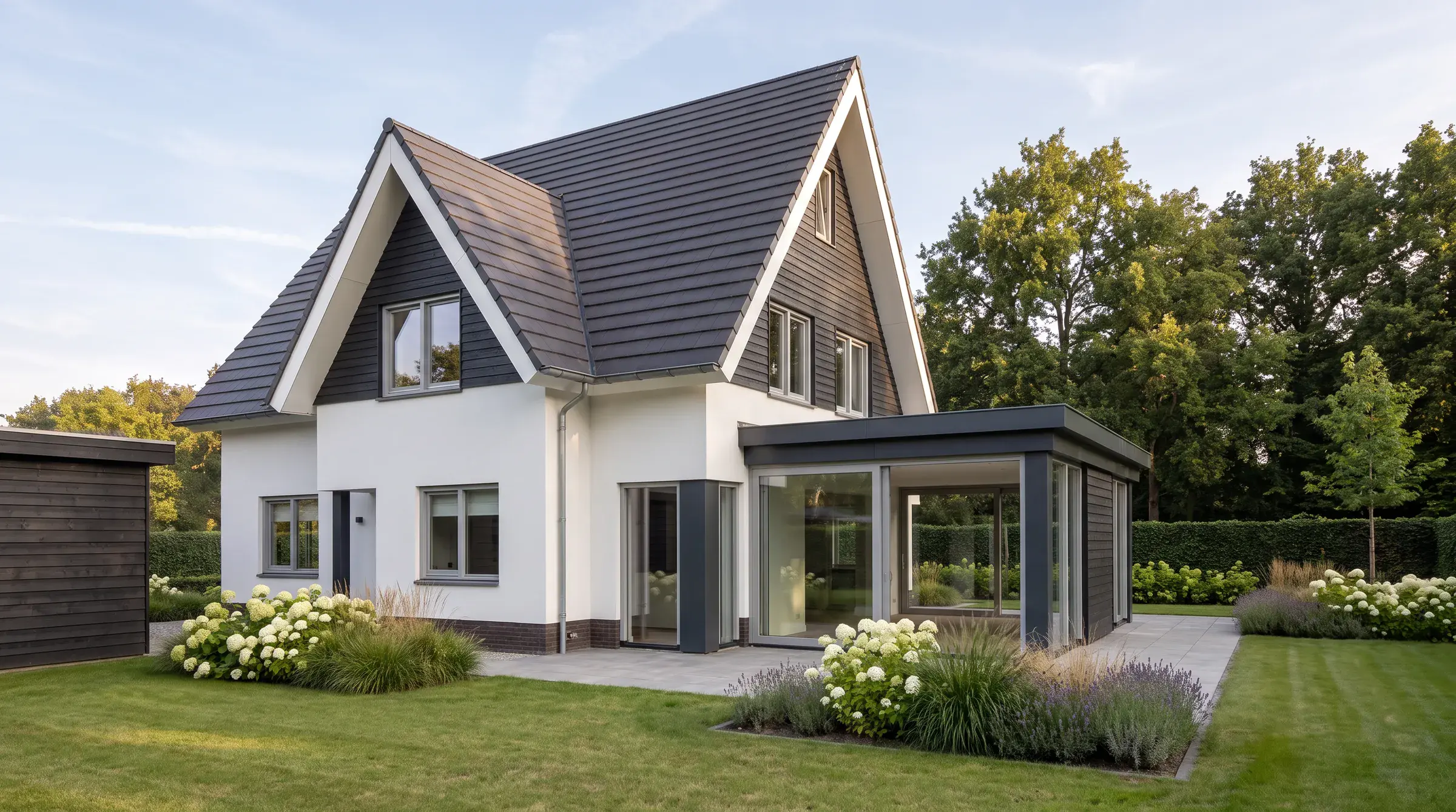 Impressie van de tuinhoek: een wit gestucte villa onder een steile donkere kap met donkere houten delen in de topgevels, rechts de glazen tuinkamer onder een plat dak